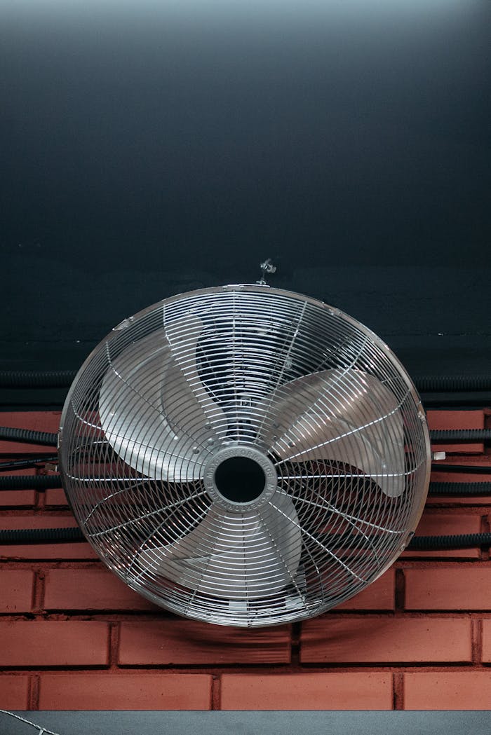 Close-up of an industrial ceiling fan with metal blades mounted on a brick wall indoors.
